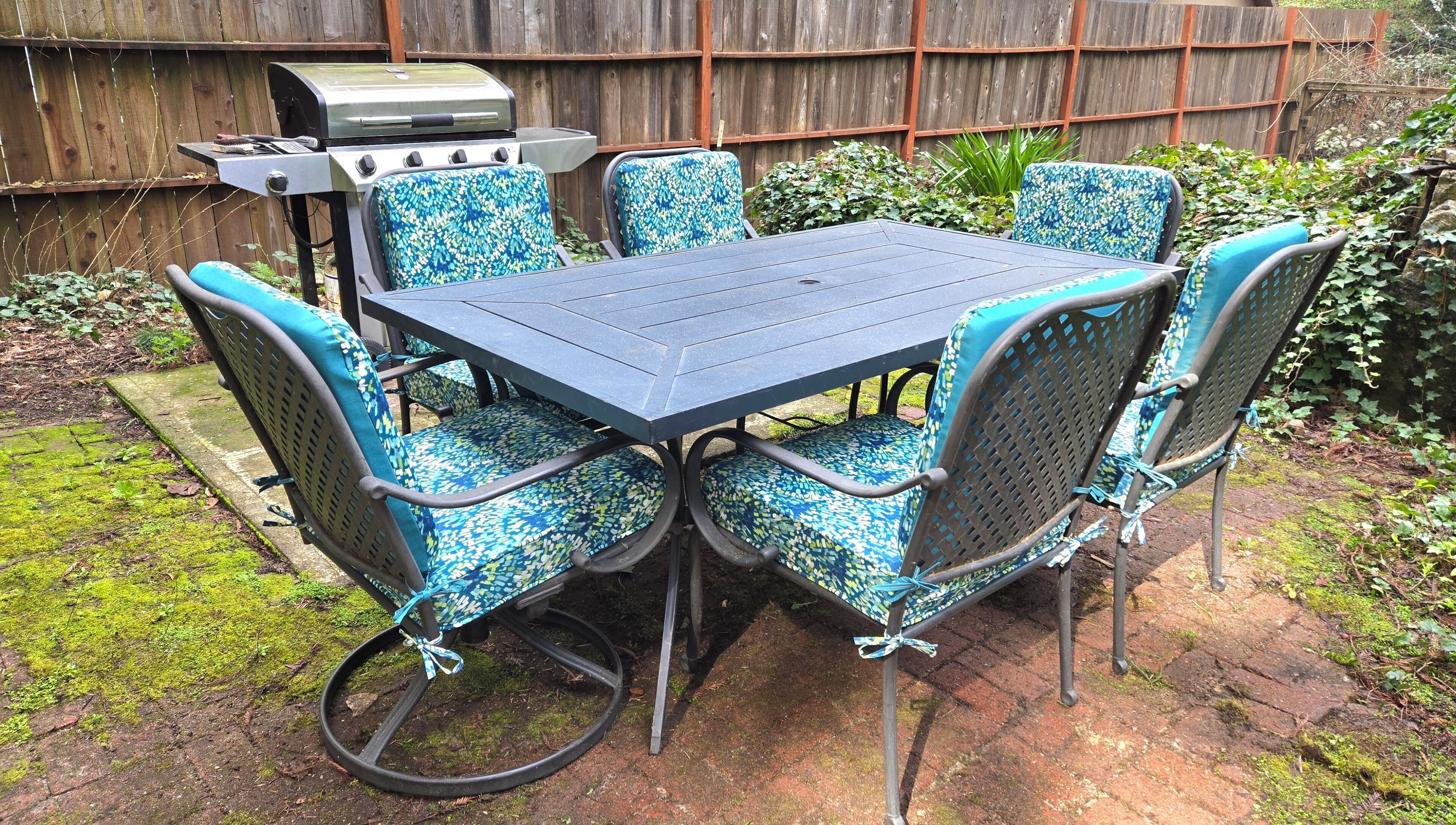 An outdoor patio with a table and chairs, a grill, and a wooden fence in the background.