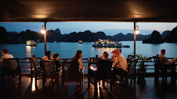 A covered outdoor seating area with wooden chairs and tables, overlooking a body of water with boats and distant mountains. - Le Journey Premium Cruise (Tuan Chau)