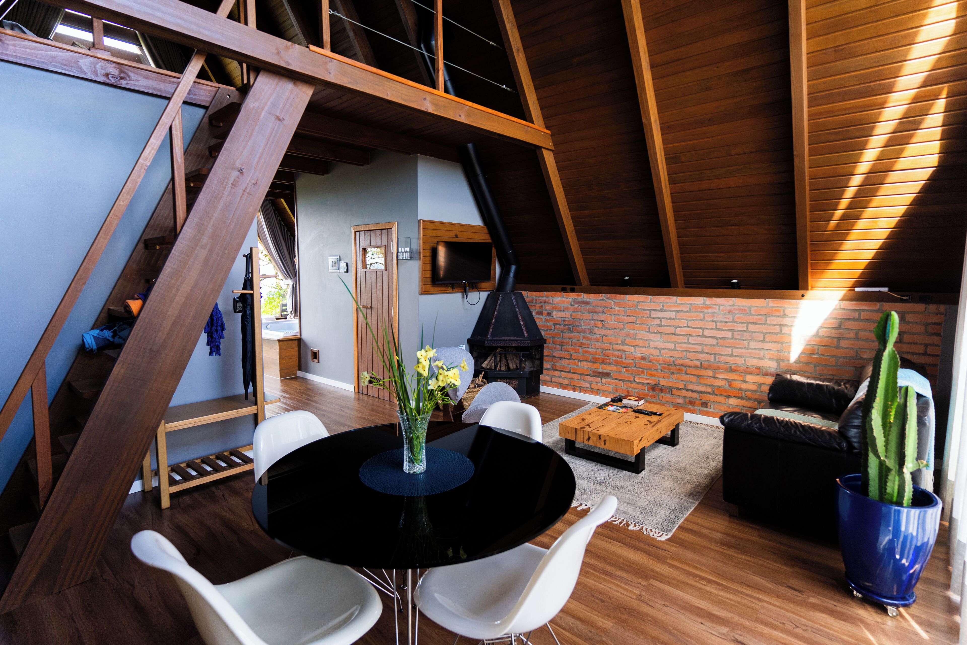 A spacious loft with a high ceiling, exposed wooden beams, a brick fireplace, and a dining area with a black table and white chairs.