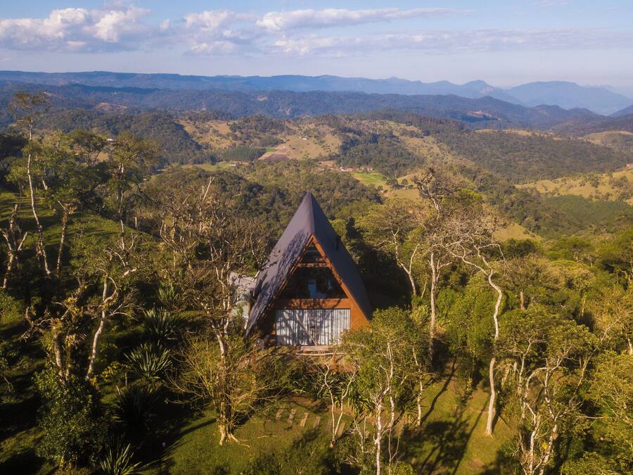 Uma cabana isolada, com telhado triangular, rodeada por floresta densa, com vista panorâmica para montanhas distantes.