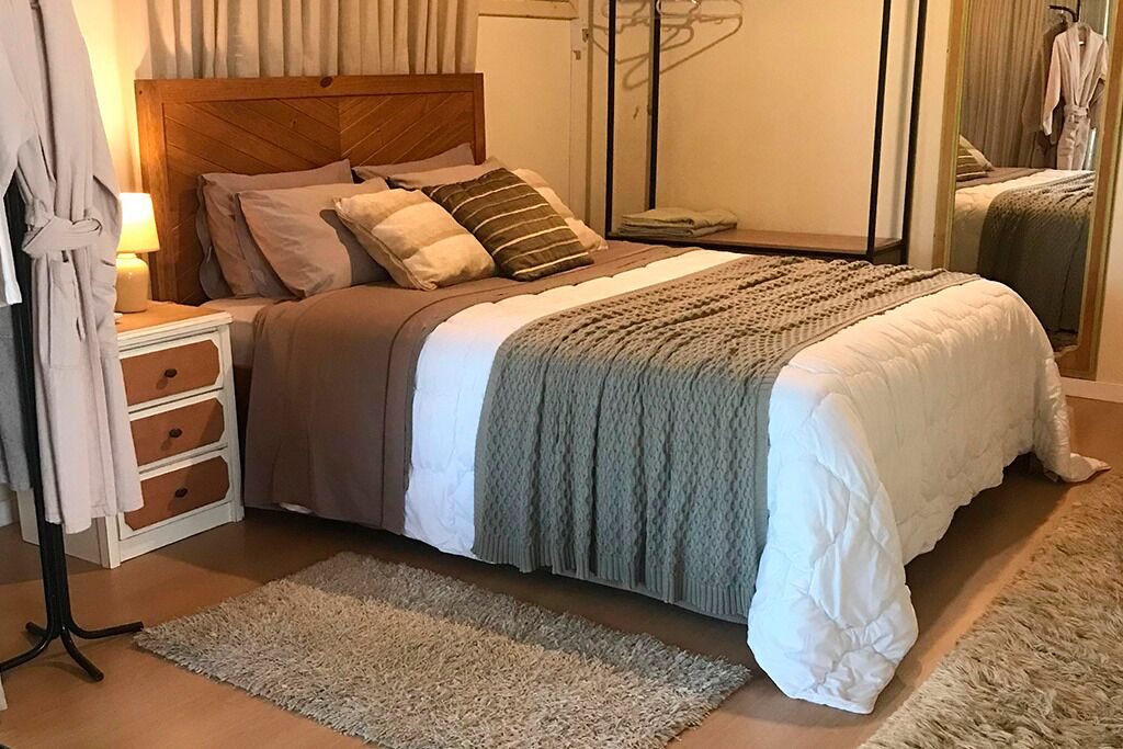 Un dormitorio ordenado con una cama grande, una mesita de noche con lámpara, un espejo y una bata colgada en un perchero.