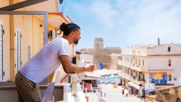 A man leaning on a balcony railing overlooking a street with buildings and a clear sky.