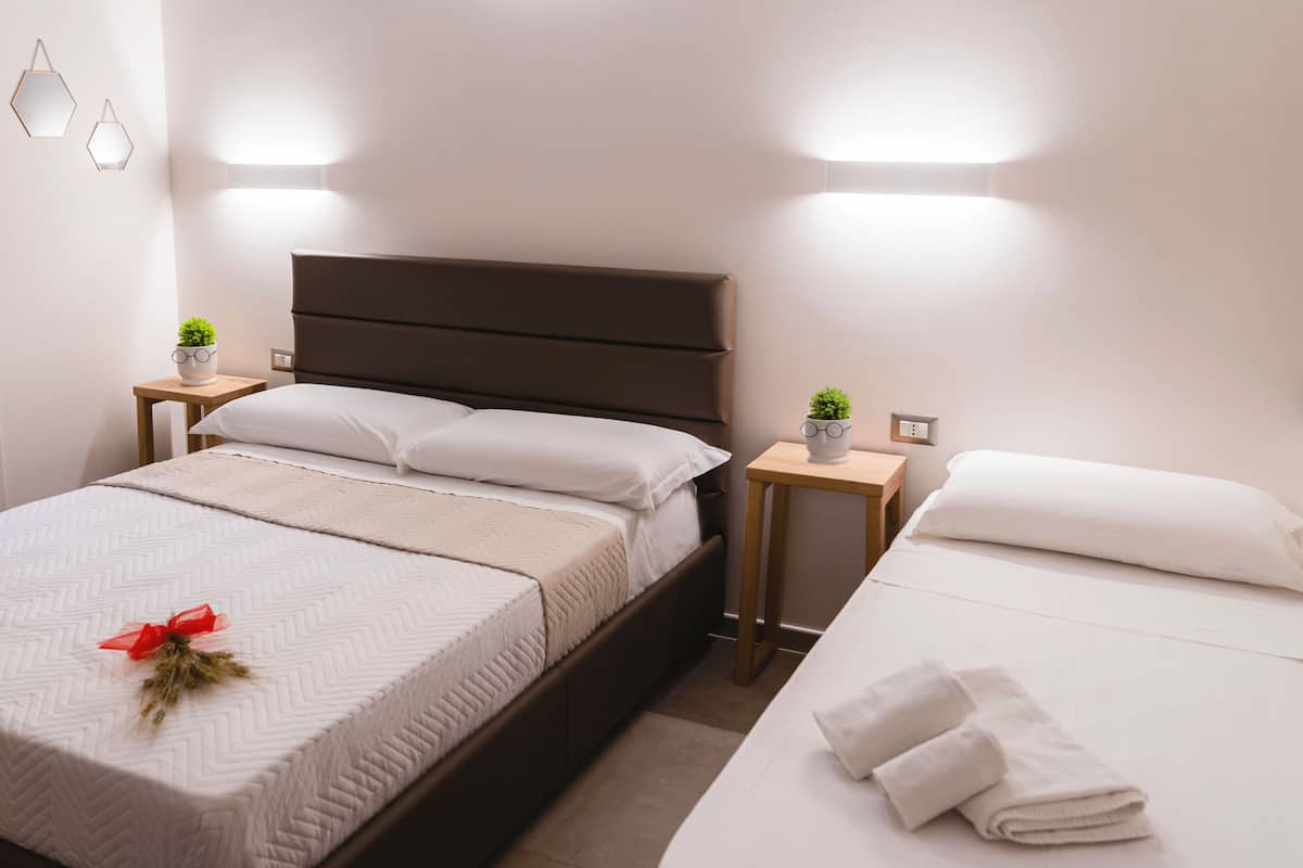 A neatly made bed with a white comforter and a red bow, a bedside table with a small plant, and a wall-mounted light fixture.