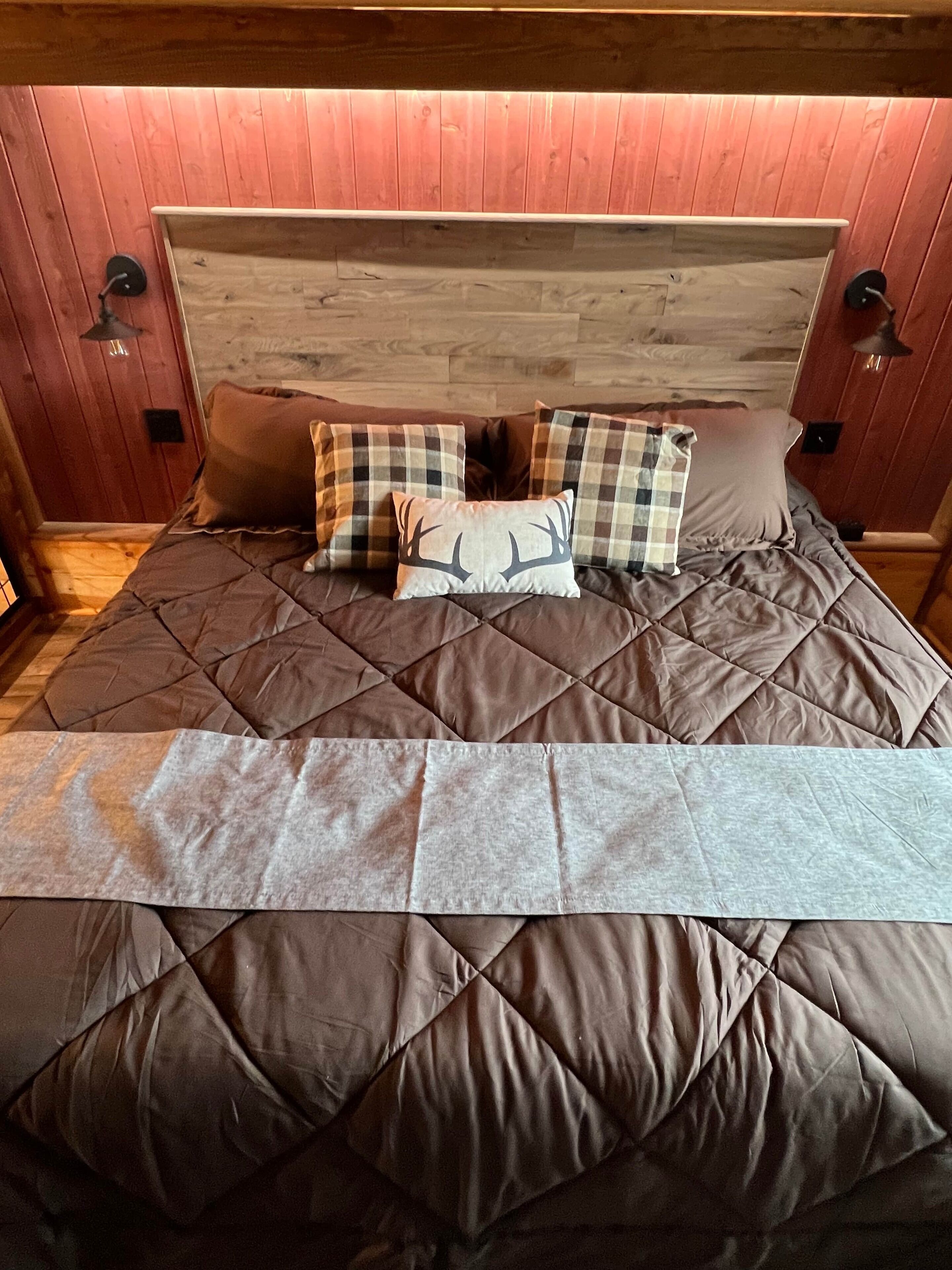 A bed with a quilted brown cover and plaid pillows, including one with a deer design, in a room with wooden walls and a wooden headboard.