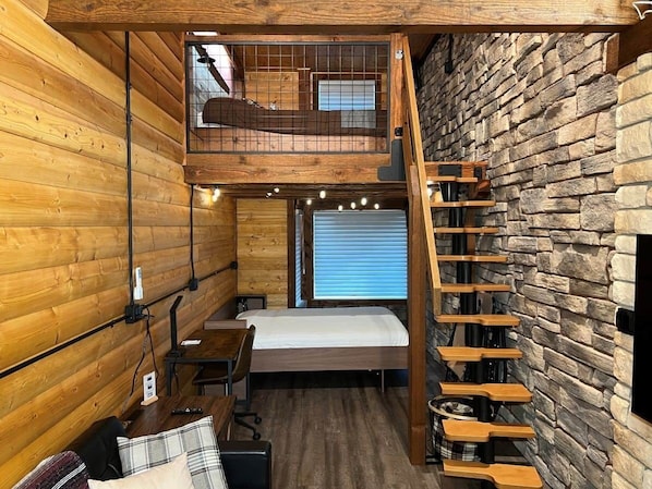 A compact room with a loft bed, a desk, and a staircase against a stone wall.