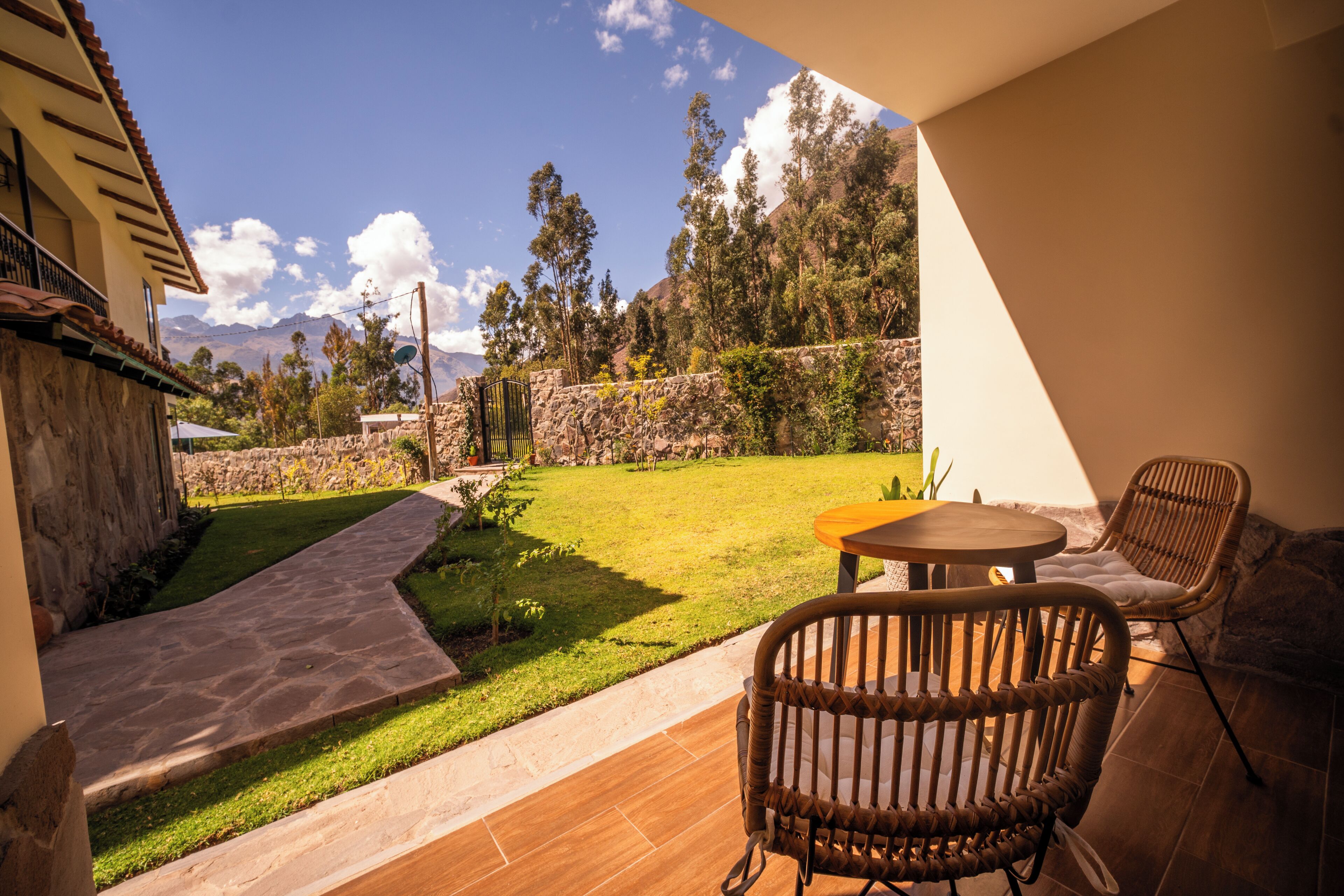 A patio with wicker chairs and a table, a stone pathway, and a view of a grassy yard with trees and a mountain in the background.