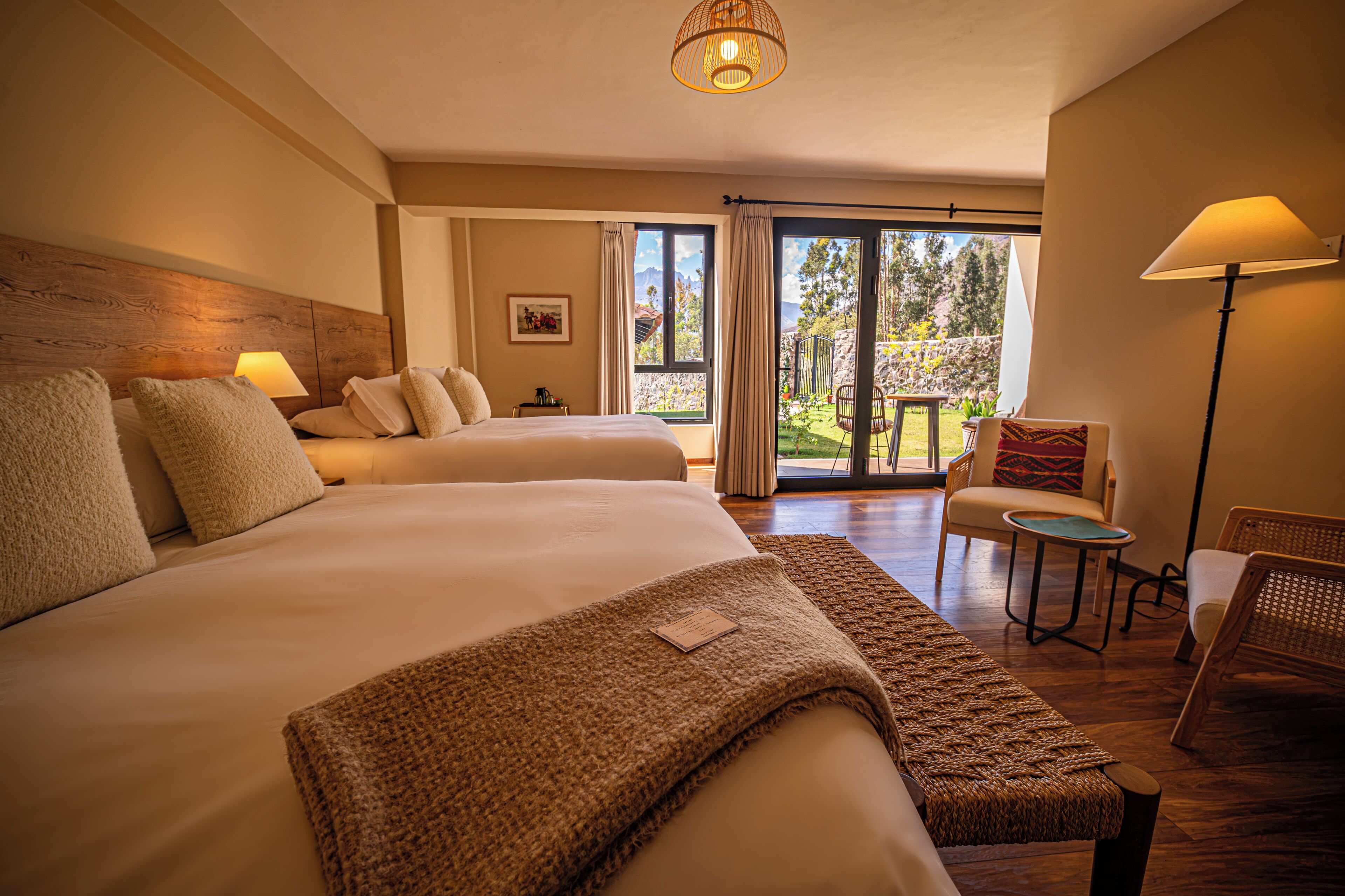 Habitación de hotel con dos camas, una zona de estar con una silla y una mesa, y un amplio ventanal corredero a un espacio exterior.