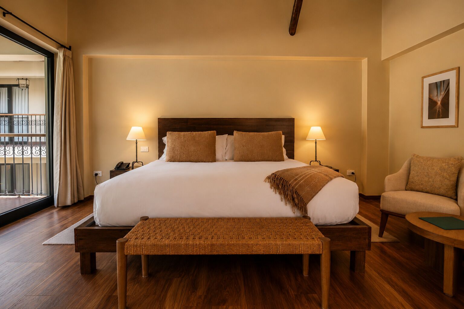Habitación de hotel con una cama grande, dos lámparas de noche, una silla, una mesita y vistas a través de una puerta de balcón.