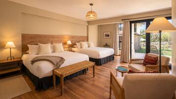 A hotel room with two beds, a sitting area with a chair and a table, a wooden floor, and a large window with curtains.