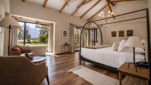 Un dormitorio con una cama grande, un sofá, una silla y una mesa. Hay ventanas con cortinas, una araña y vistas al exterior.