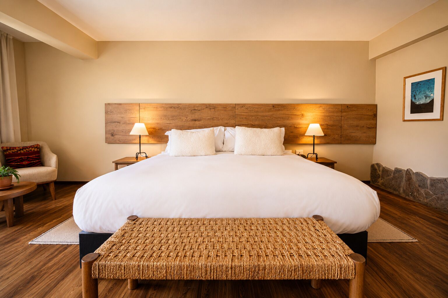 Una cama grande con cabecera de madera, dos lámparas de noche, un banco tejido, una mesita con una planta y un cuadro en la pared.