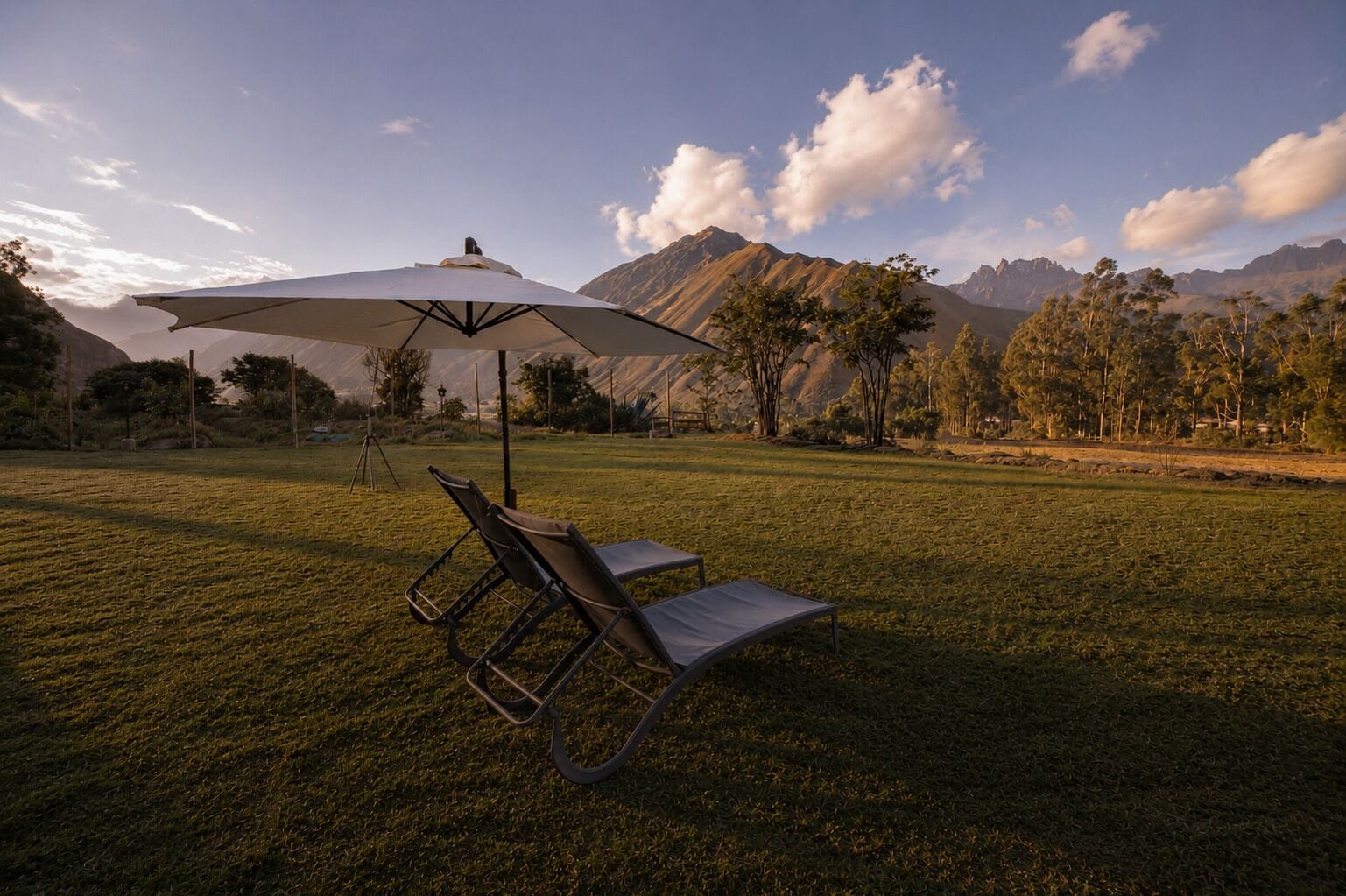 Un espacio al aire libre con césped, una silla de descanso y una sombrilla, rodeado de montañas y árboles.