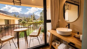 Un balcón con mesa y sillas, un baño con lavamanos y espejo grandes, y vistas a montañas y vegetación.