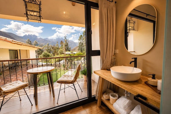 A balcony with a table and chairs, a bathroom with a large sink and mirror, and a view of mountains and greenery.