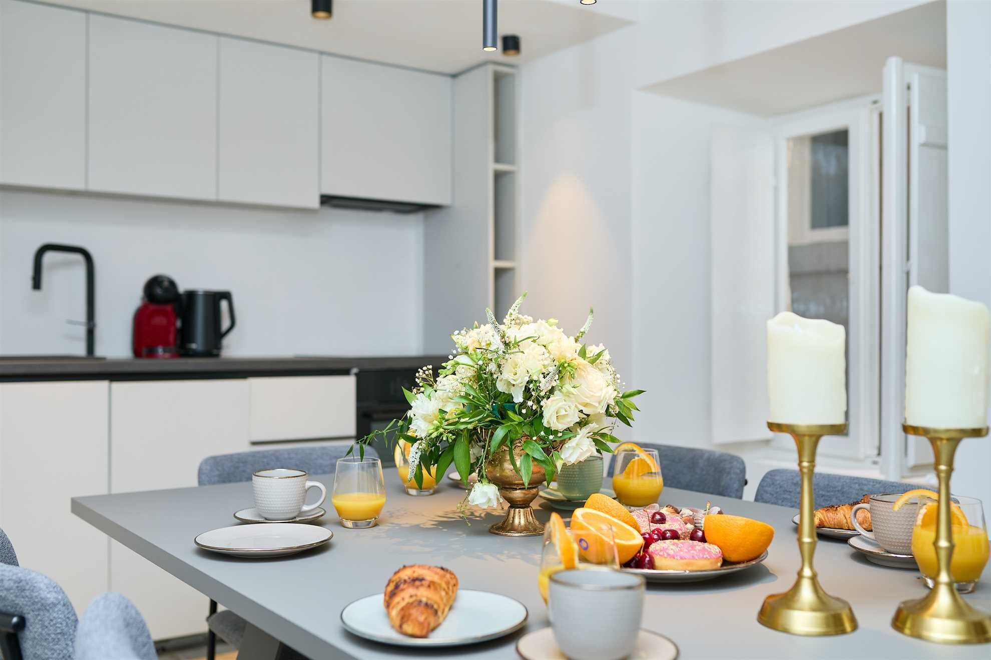 A modern kitchen with white cabinets, a coffee maker, and a dining table set with breakfast items including croissants, fruit, and a vase of flowers.