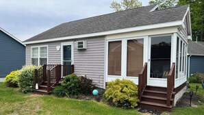 A single-story house with a covered porch, a front door, and a small front yard with bushes and a lawn. - Charming Cottage for Rent at Beach Dreams Cottages, Wells, Maine! Unit #229 (Wells)