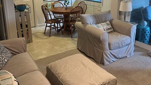 A living room with a beige sofa, a matching ottoman, and a dining area with a wooden table and chairs. - Charming Cottage for Rent at Beach Dreams Cottages, Wells, Maine! Unit #229 (Wells)