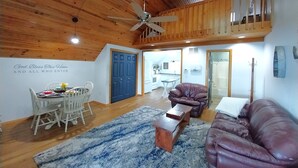 A spacious living area with a high ceiling, wooden beams, a ceiling fan, a dining table with chairs, a brown leather sofa, and a kitchen in the background. - Kansas Lake House With Private Boat Dock (Anthony)