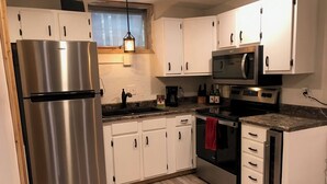 A modern kitchen with white cabinets, stainless steel appliances, a wooden floor, and a window with a hanging light fixture. - Cozy Condo (Cass)