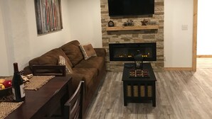 A living room with a stone fireplace, a flat-screen TV mounted above it, a brown sofa with cushions, and a small table with a bottle and glasses. - Cozy Condo (Cass)
