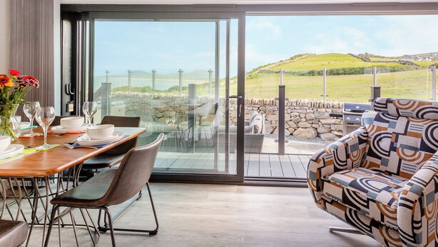 A dining area with a table set for two, a sofa, and a view of a grassy landscape through large glass doors. - 3 bedroom accommodation in Llandanwg (Llandanwg)