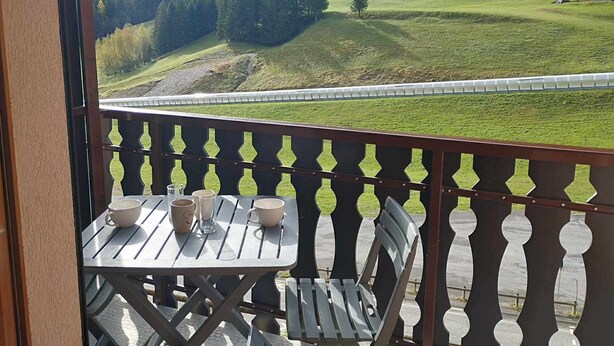 Un balcon avec une table et des chaises, donnant sur un paysage verdoyant avec une colline boisée et une voie ferrée.