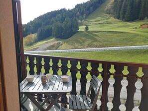 Un balcón con mesa y sillas, con vistas a un paisaje verde que incluye una colina boscosa y una vía de tren.