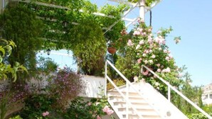 Un escalier blanc, agrémenté d’une balustrade blanche, mène à une pergola blanche recouverte de lianes vertes et de fleurs roses.