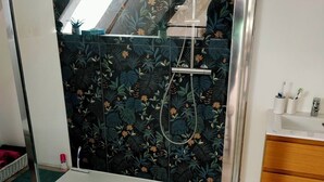 Ducha con cerramiento de vidrio, un salpicadero de azulejos con motivos florales, suelo de madera y un lavamanos pequeño con artículos de aseo.