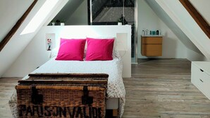 Un dormitorio tipo loft con techo inclinado, cama con almohadas rosas, un cofre de mimbre y un pequeño armario de madera.