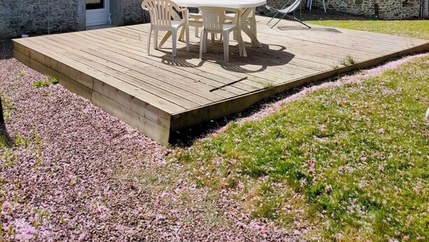 Una casa de piedra con terraza de madera, una mesa y sillas, y un pequeño jardín con flores rosadas.