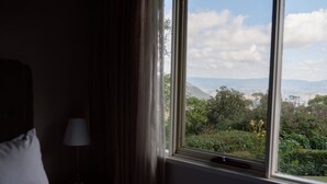 A hotel room with a bed, a nightstand, a lamp, and a window offering a view of greenery and distant mountains. - Yamacilla House: Spectacular Views in Blackheath! (Blackheath)