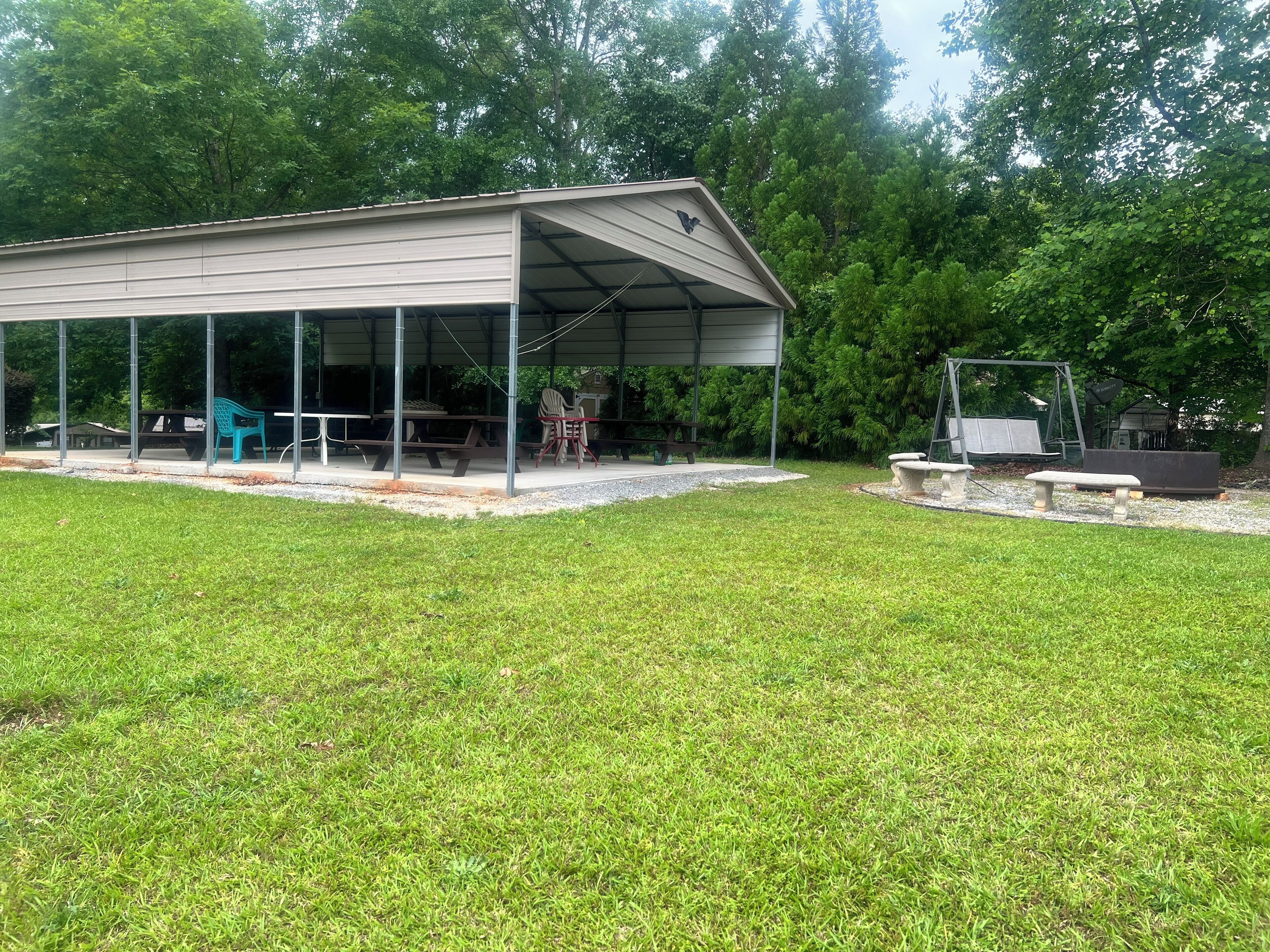 A covered outdoor area with picnic tables and chairs, surrounded by a grassy field and trees.