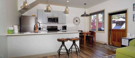 A compact kitchen with a microwave, stove, and bar stools, a dining area with a wooden table and chairs, and a large window with a view.