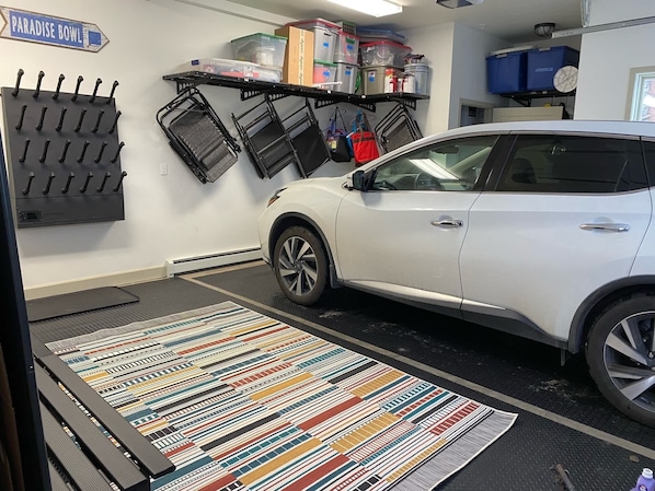 Un VUS blanc est garé dans un garage où l’on trouve un tapis à rayures colorées, un présentoir métallique avec des crochets, ainsi que des étagères de rangement avec des bacs et des contenants.
