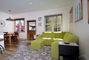 A living room with a green sofa, a dining table with chairs, a clock, and large windows. - Horseshoe Haven, Immaculate 2BD/2BA, Pet friendly! (Mt. Crested Butte)