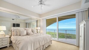 Une chambre dotée d’un grand lit, d’un ventilateur de plafond, d’une table de chevet avec une lampe, d’une commode et d’une grande fenêtre donnant sur un balcon.