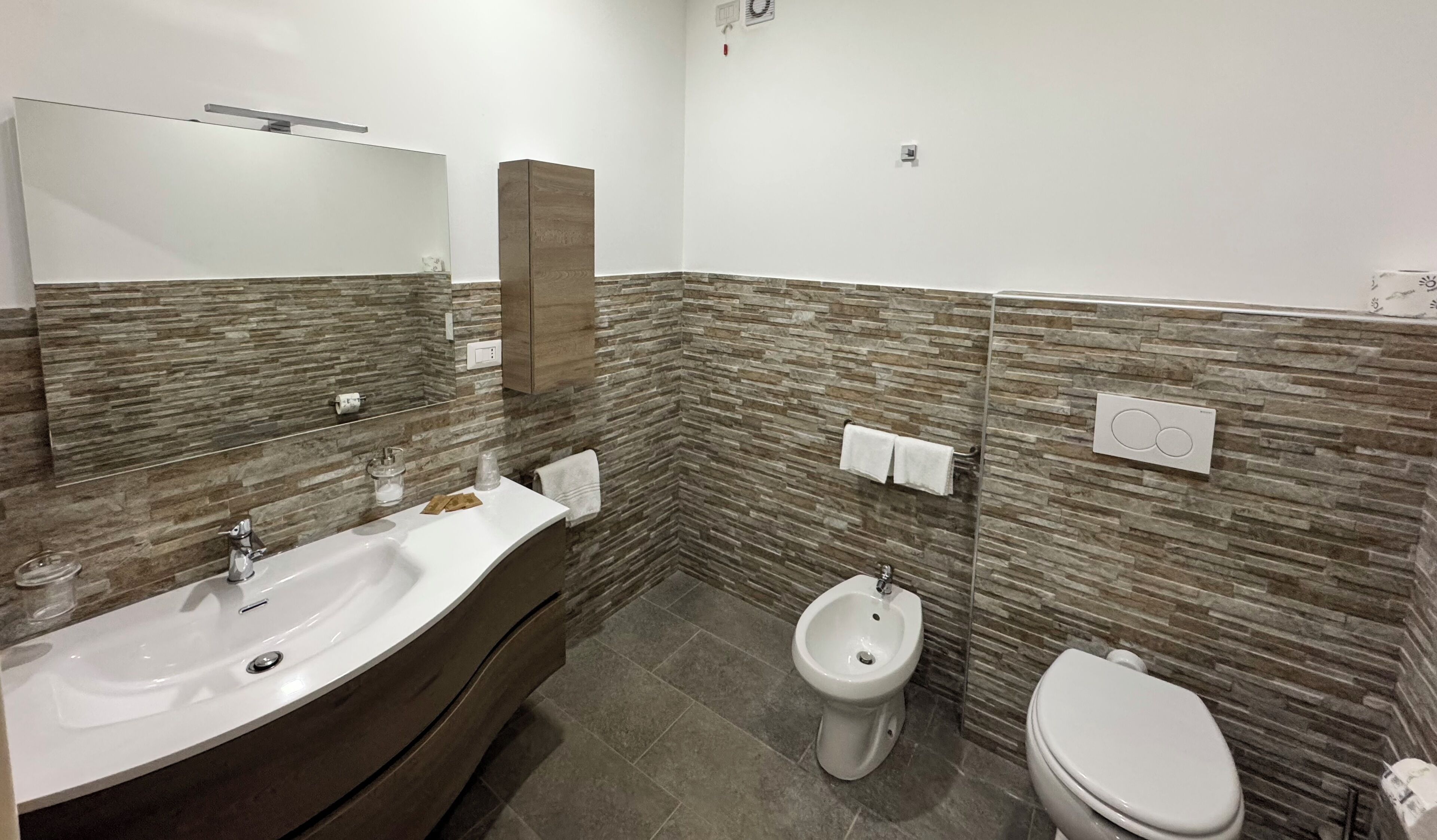 Un baño con un lavamanos grande, un inodoro y un bidé. Las paredes están cubiertas de azulejos con un diseño que imita la piedra.
