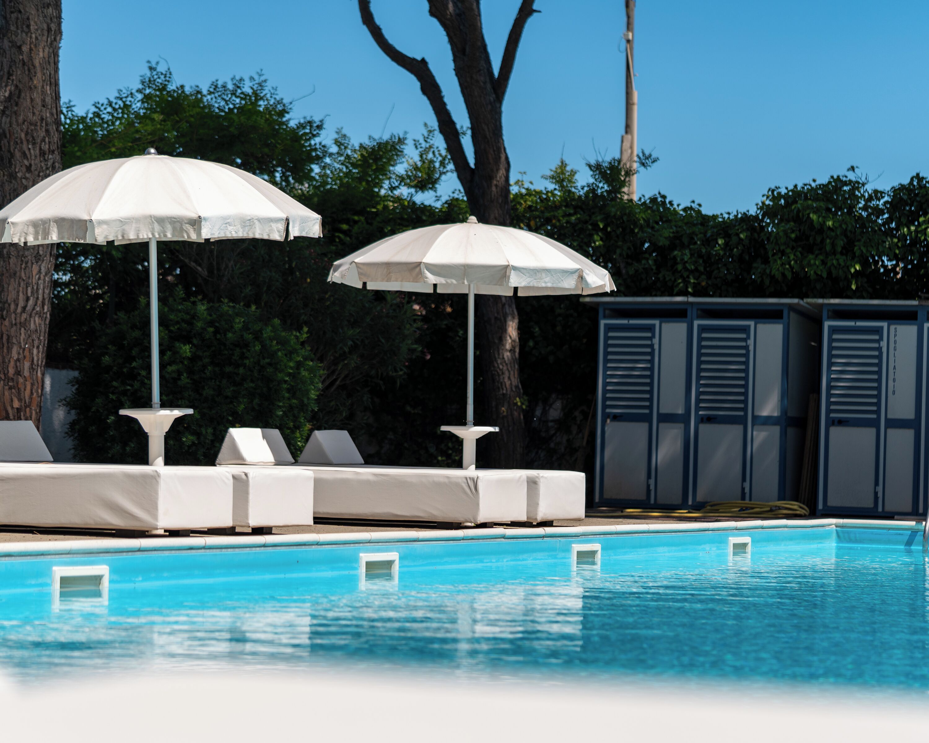 Area piscina con poltrone da relax bianche e ombrelloni, circondata da alberi e un edificio con persiane metalliche.
