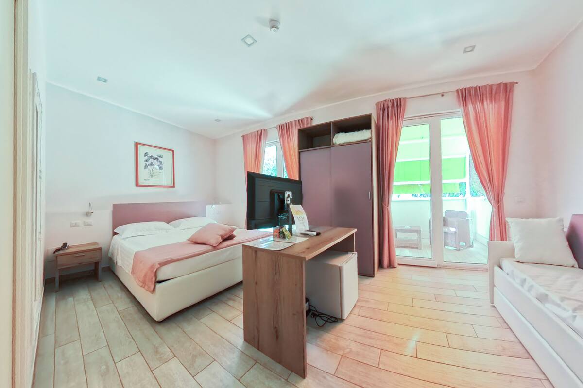 A hotel room with a bed, a desk, a television, and a balcony with curtains.