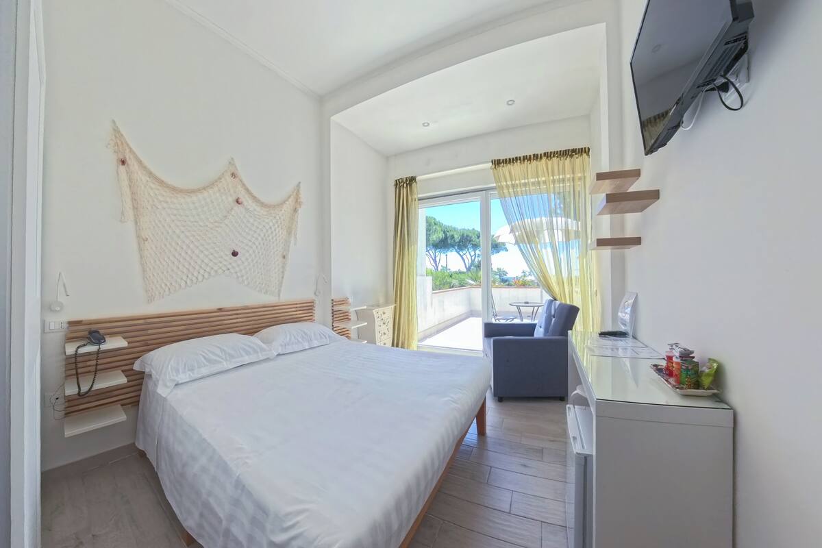 A hotel room with a bed, a desk with a TV, a chair, and a balcony with curtains.