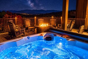 A hot tub with a fire feature, surrounded by wooden chairs and a railing, set against a night sky with stars and a mountain backdrop.