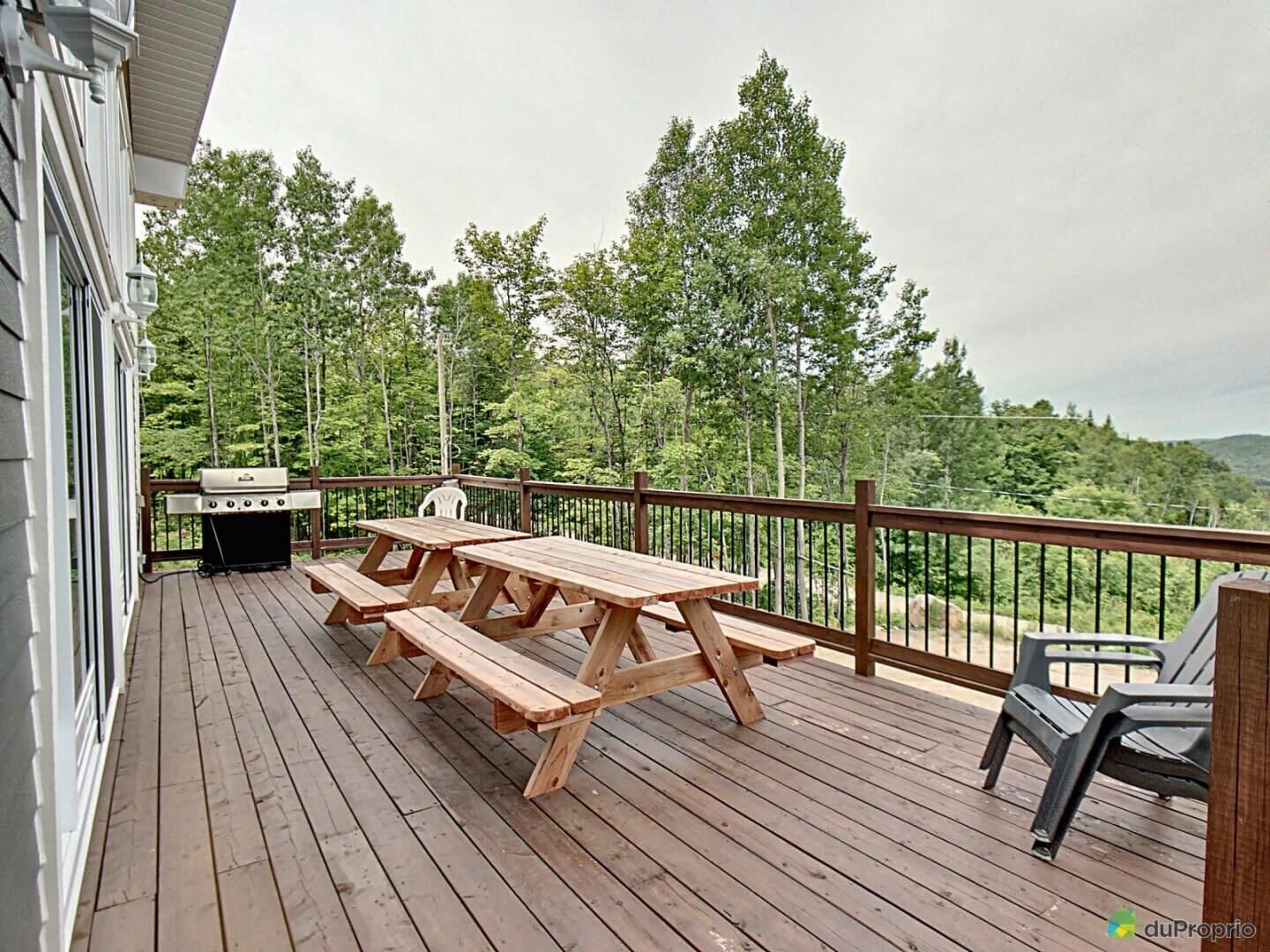 A wooden deck with picnic tables and a grill, surrounded by trees and a view of the landscape.