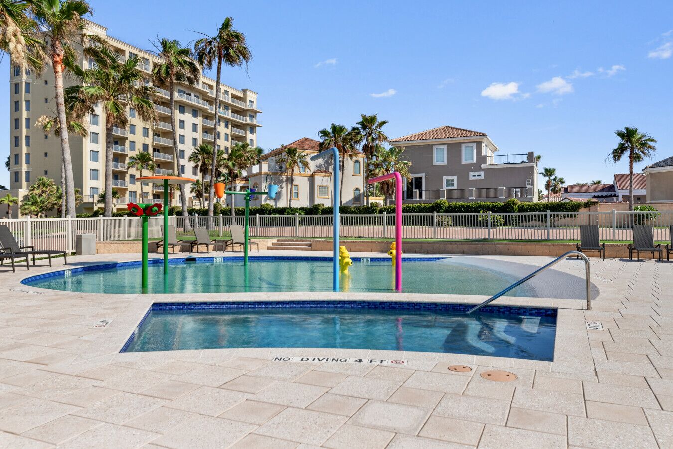 A swimming pool with a no diving sign, surrounded by palm trees and lounge chairs, with buildings in the background.