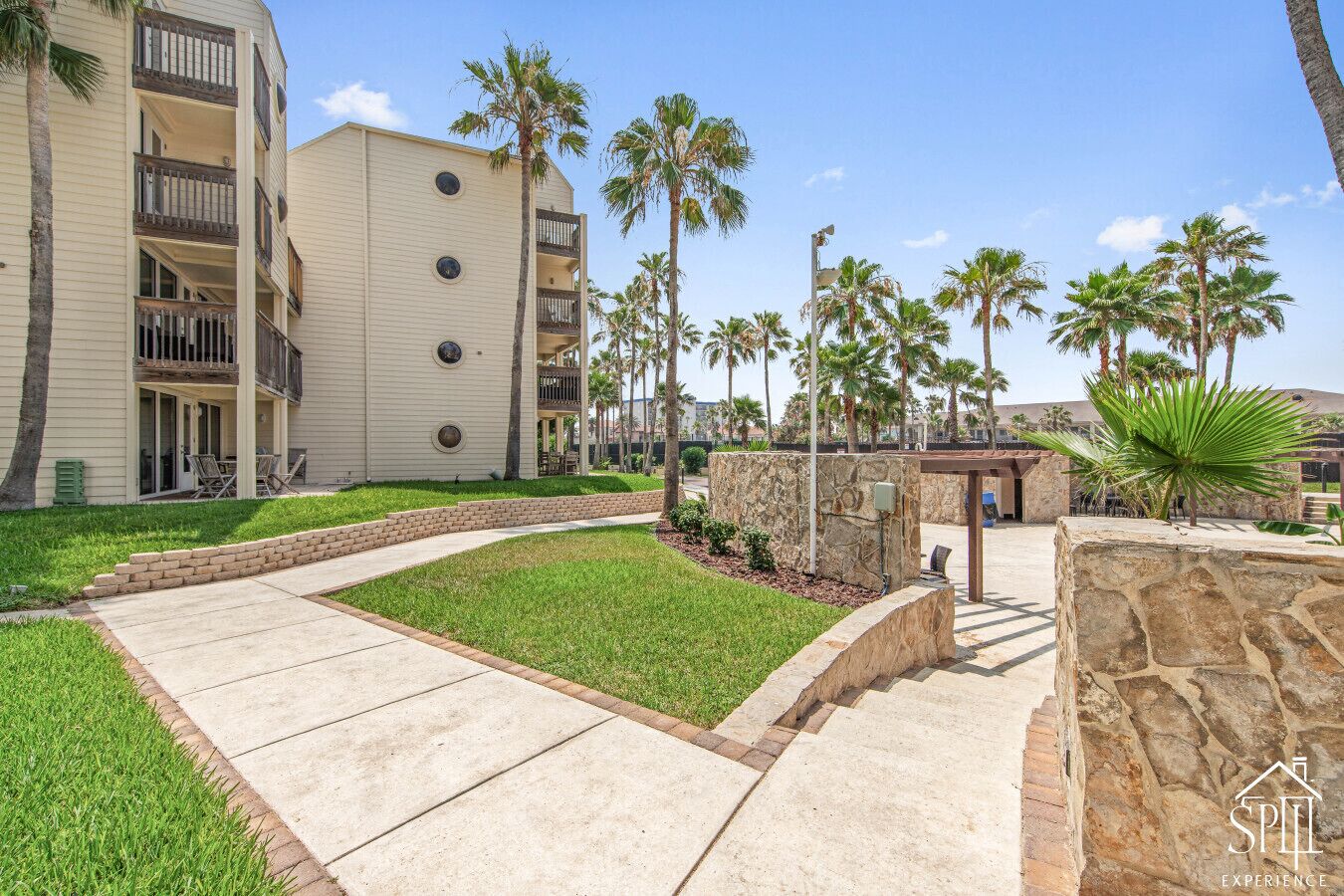 A multi-story hotel with a covered outdoor area, palm trees, and a stone wall.