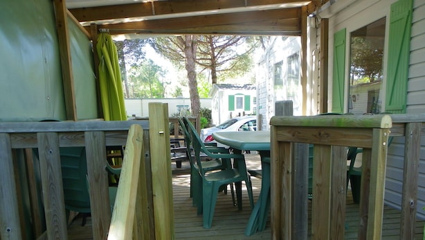 A wooden deck with a table and chairs, a covered area with a door, and a view of a white house with green shutters in the background. - Découvrez le Camping Bois Soleil Dans le Confort de Notre Mobil Home ! (Saint-Georges-de-Didonne)
