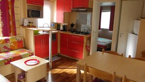 A compact kitchen with red cabinets, a microwave, and a small dining area with a table and chairs. - Découvrez le Camping Bois Soleil Dans le Confort de Notre Mobil Home ! (Saint-Georges-de-Didonne)