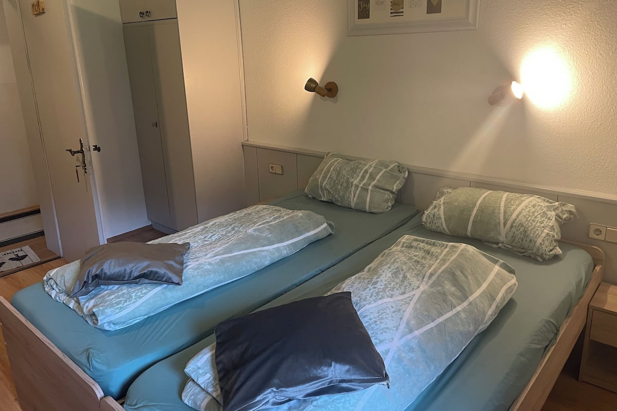 A room with two single beds, each with a pillow and a blanket. There is a wardrobe with a mirror and a door handle.