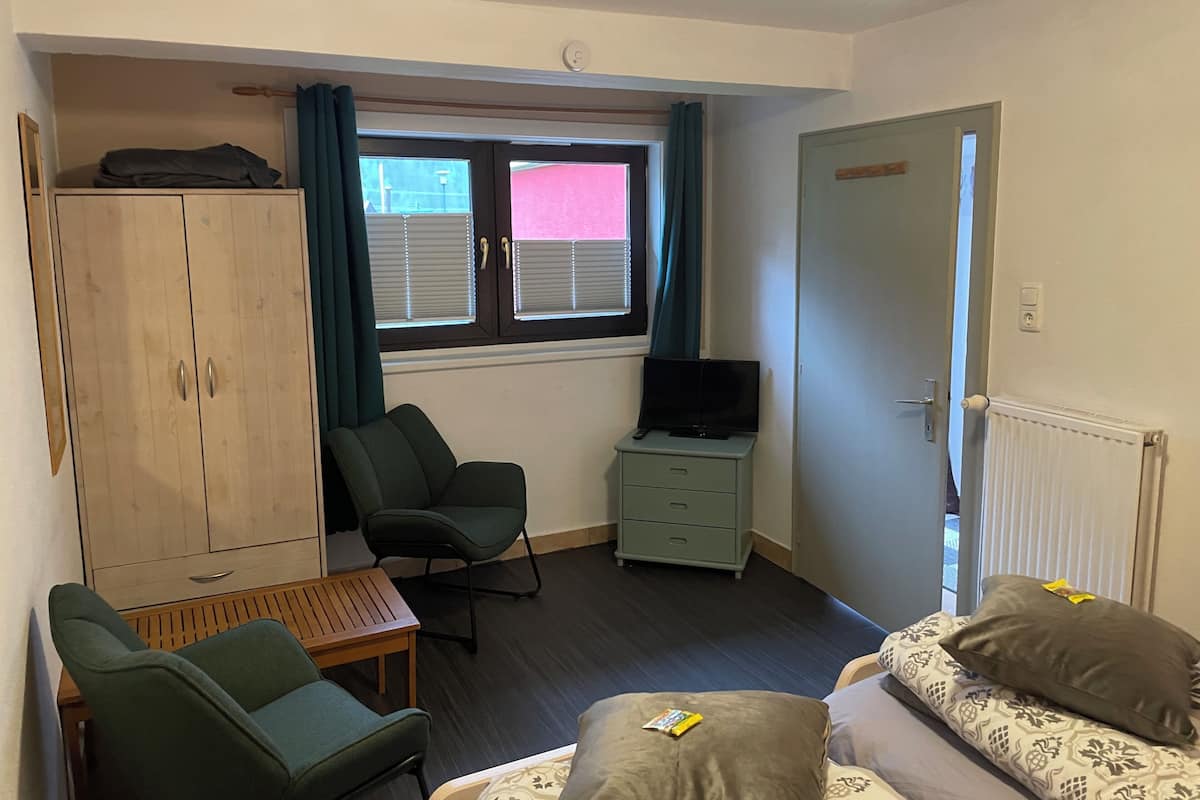 A compact hotel room with a single bed, a desk, a chair, a wardrobe, a television, and a window with blinds.