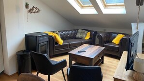A cozy living room with a leather sofa, a coffee table, and a fireplace. There are skylights on the ceiling, a hanging plant, and a TV on a stand. - Molltaler Appartements  (Stall)
